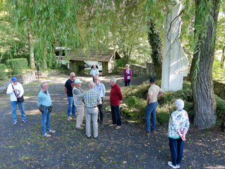 Gemischte Gruppe älterer Menschen im Freien, umgeben von Bäumen, Steinmauern und einem weißen Denkmal, mit Bus im Hintergrund.