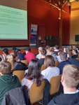 Ein Redner präsentiert vor einem jungen Publikum in einem Vortragssaal; deutscher Text über Meinungsfreiheit auf dem Bildschirm.