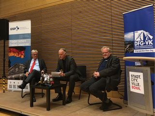 Drei Männer auf einer Bühne bei einer Podiumsdiskussion, umrahmt von Bannern einer Sicherheitspolitik- und Friedensgesellschaft.