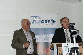 Zwei Männer präsentieren bei einer 70-Jahr-Feier der GSP (Gesellschaft für Sicherheitspolitik), einer mit Mikro, der andere am Rednerpult.