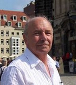 Nahaufnahme eines älteren Mannes in einem weißen Hemd, mit historischen Gebäuden und Personen im unscharfen Hintergrund.