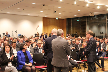 Zuschauer und Redner bei einer Konferenz in einem großen, hellen Raum; die meisten Teilnehmer sitzen.