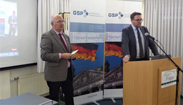 Zwei Männer bei einer GSP-Veranstaltung, einer am Rednerpult, mit Bannern der deutschen Flagge und des Reichstagsgebäudes.