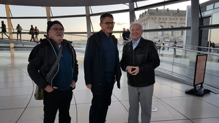 Drei lächelnde Männer in der Reichstagskuppel, mit der historischen Reichstagsfassade im Hintergrund sichtbar.