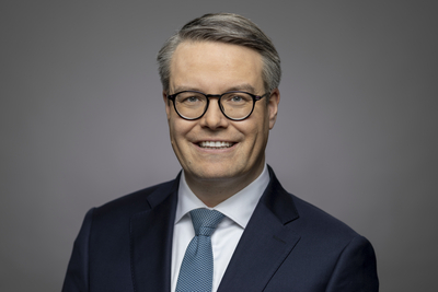 Kopfaufnahme eines lächelnden Mannes mit grauem Haar, Brille, dunklem Anzug, weißem Hemd und blauer Krawatte vor grauem Hintergrund.