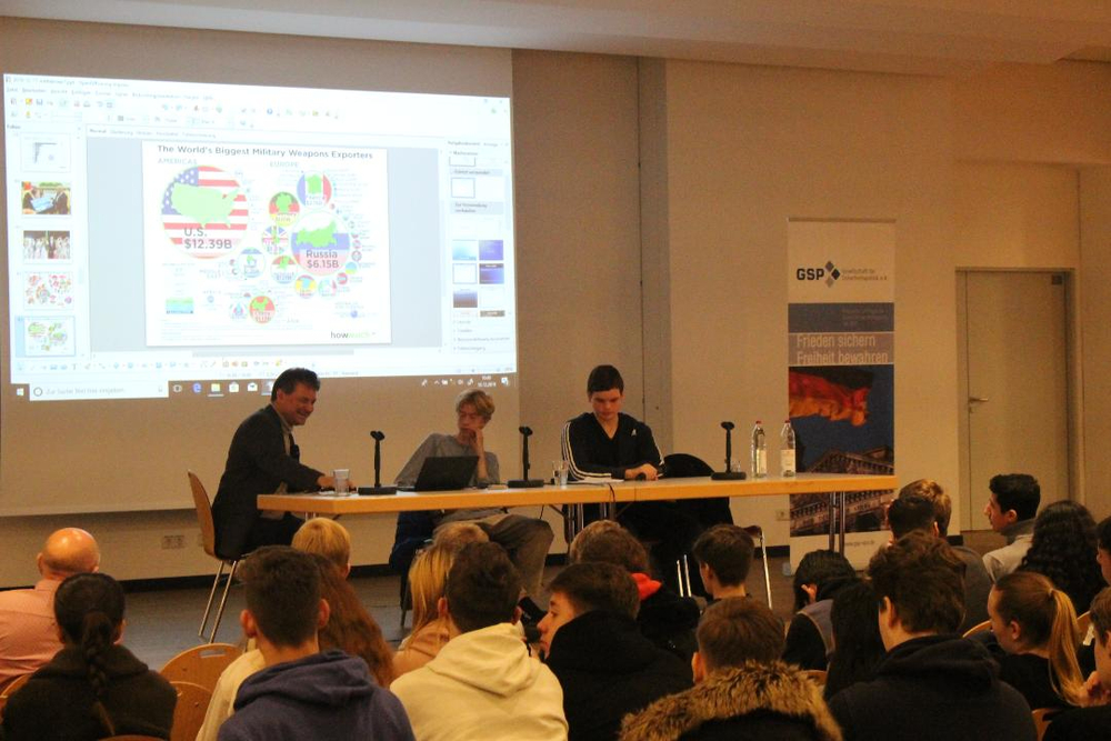 Podiumsdiskussion mit Referenten und Publikum, eine Leinwand zeigt Waffenexportdaten. Ein GSP-Banner steht im Hintergrund.