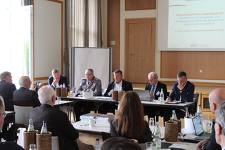 Männer an einem Konferenztisch, darunter ein Mann in Militäruniform, bei einer Präsentation über Sicherheitspolitik.
