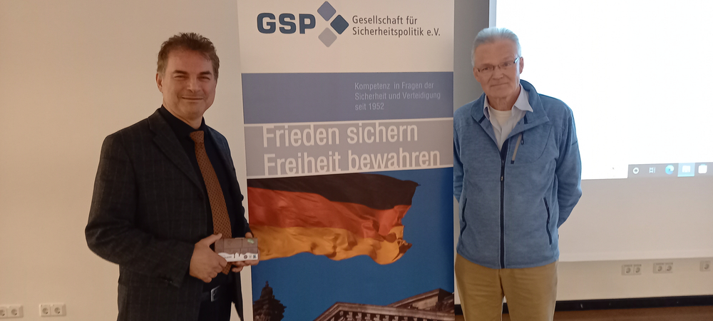 Zwei Männer, einer mit kleinem Paket, lächeln vor einem GSP-Banner mit deutscher Flagge.
