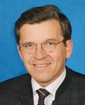 Porträt eines lächelnden Mannes mit dunklen Haaren, Brille, dunkler Jacke, gestreiftem Hemd und Krawatte vor blauem Hintergrund.