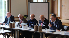 Fünf Männer nehmen an einem Konferenztisch teil, einer spricht ins Mikrofon, während andere Notizen machen.
