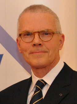 Mann mit grauem Haar, Brille, Anzug und gestreifter Krawatte lächelt vor einem weißen Hintergrund mit blauem Logo.