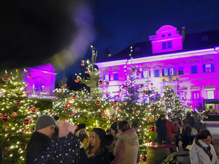 Eine festliche Nachtszene mit Menschen zwischen beleuchteten Weihnachtsbäumen, roten Kugeln und magentafarbenen Gebäuden.