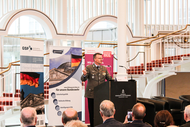 Militäroffizier Wüstner spricht vor Publikum bei einer Konferenz. Im Hintergrund sind politische Banner und die deutsche Flagge zu sehen.