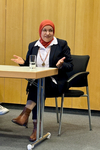 Frau mit rotem Hijab, Brille und schwarzem Blazer sitzt an einem Tisch, gestikuliert, mit einem Glas Wasser.
