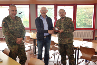 Drei Männer, zwei in deutscher Uniform, ein Zivilist. Der Zivilist schüttelt einem Soldaten die Hand, der eine Auszeichnung entgegennimmt.