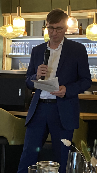 Ein Mann im blauen Anzug mit Brille spricht in ein Mikrofon, Papiere haltend, in einem Restaurant oder einer Bar mit Lichtern.