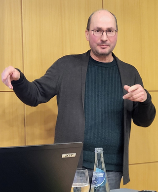 Ein Mann mit Brille in dunklem Pullover und Strickjacke gestikuliert bei einem Vortrag, Laptop und Getränke vor ihm.