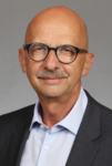 Lächelnder kahler Mann mit Brille und Schnurrbart, in blauem Hemd und grauem Sakko.