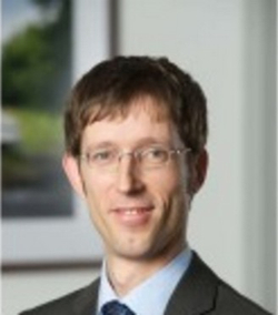 Professionelles Porträt eines Mannes mit kurzen braunen Haaren, Brille und Anzug, leicht lächelnd.