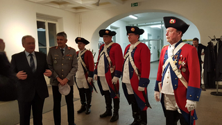 Männergruppe: einige in historischen roten Militärmänteln & schwarzen Hüten, andere in modernem Anzug & Militäruniform, drinnen.