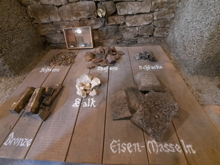 Eine Ausstellung zeigt verschiedene Materialien: Bronzebarren, Eisenmasseln, Schlacke, Bohnerz, Stuferz und Kalk auf Holz.