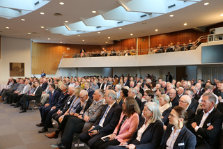 Großes Publikum, darunter Militärpersonal und Zivilisten, bei einer Konferenz in einem modernen Auditorium mit Balkon.