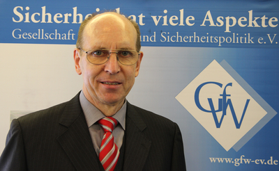 Porträt eines lächelnden Mannes mit Brille, Anzug und gestreifter Krawatte vor einem blauen Banner mit Text über Sicherheitspolitik.