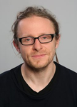 Porträt eines Mannes mit hellbraunem Haar im Dutt, schwarzer Brille, leichtem Bart und schwarzem Hemd, leicht lächelnd.