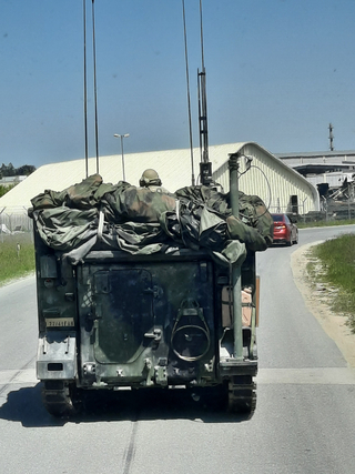 Rückseite eines militärischen Kettenfahrzeugs mit Soldat im Helm, Antennen und Ausrüstung fährt auf Straße an Gebäuden vorbei.