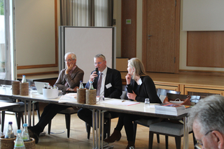 Drei Personen an einem Konferenztisch. Ein Mann spricht in ein Mikrofon, flankiert von zwei aufmerksam zuhörenden Teilnehmern.