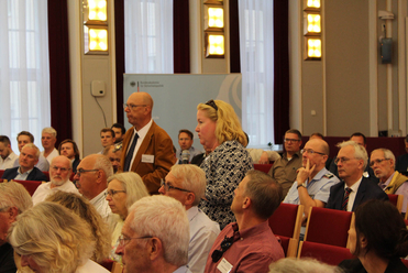 Eine Frau spricht vor einem Publikum aus mostly älteren Männern und Frauen bei einer Konferenz oder Veranstaltung.