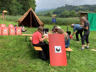 Menschen essen im Freien bei einem Römer-Reenactment-Event mit Zelt, Schilden und Bannern auf einer Wiese.