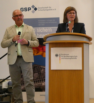 Mann mit Mikrofon und Frau am Rednerpult bei einer Konferenz mit Logos von GSP und Bundesamt für Bevölkerungsschutz.