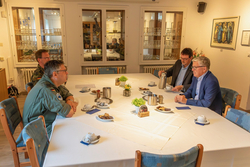 Vier Männer, zwei in Uniform und zwei in Anzügen, sitzen an einem großen weißen Tisch mit Kaffee und Snacks und unterhalten sich.