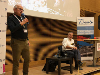 Ein Mann mit Brille spricht in ein Mikrofon bei einer Konferenz, mit Veranstaltungsbannern und einem Zuhörer im Hintergrund.