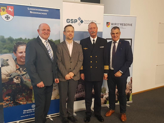 Vier Männer, darunter ein Marineoffizier, stehen lächelnd vor Bannern der Bundeswehr, Landeskommando Niedersachsen, GSP und Wir. Sind. Die. Reserve.