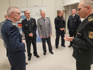 Männer in deutschen Militär- und Feuerwehruniformen im Gespräch, mit Bannern der Deutschen Atlantischen Gesellschaft.