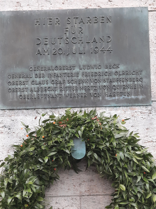 Eine Bronzetafel erinnert an die Verschwörer des 20. Juli 1944, darunter ein grüner Kranz mit orangefarbenen Beeren.