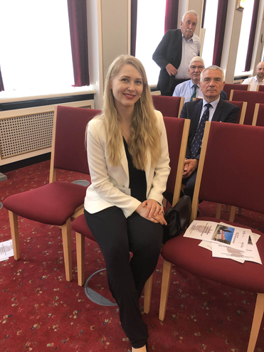 Eine lächelnde Frau mit langen blonden Haaren in einem weißen Blazer sitzt in einem Konferenzraum mit roten Stühlen.