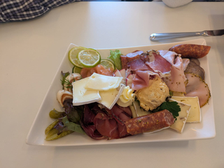 Platte mit verschiedenen Wurstwaren, Käse, Lachs, Limetten, Essiggurken und einem Aufstrich.