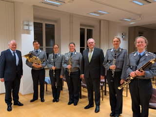 Sieben Personen: zwei in Anzügen, fünf in Uniformen halten Musikinstrumente (Horn, Oboe, Klarinetten, Fagott) und posieren.
