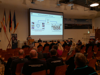 Ein Soldat präsentiert auf einer Konferenz zum OPLAN DEU vor einem sitzenden Publikum; Flaggen sind im Hintergrund.