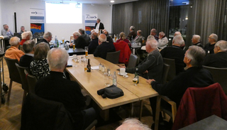 Eine Gruppe überwiegend älterer Männer sitzt an Tischen und hört einem Sprecher bei einer GSP-Veranstaltung zu.