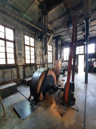 Innenansicht einer alten Industriewerkstatt mit großen, riemengetriebenen Maschinen, schweren Ketten und hellen Fenstern.