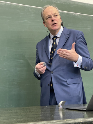 Ein Mann im blauen Anzug, mit Brille und Headset-Mikrofon, spricht und gestikuliert vor einer grünen Tafel.