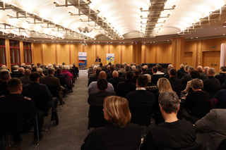 Zuschauer in einem Konferenzraum lauschen einem Redner an einem Pult.