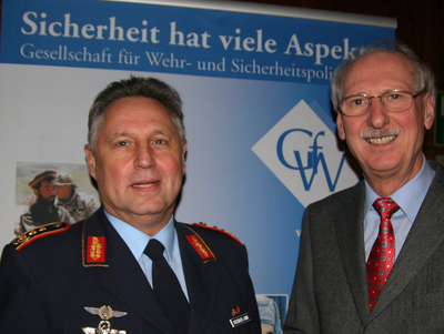 Zwei Männer, einer in Uniform, der andere im Anzug, lächeln vor einem Banner der Gesellschaft für Wehr- und Sicherheitspolitik.