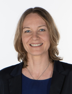 Lachende Frau mit schulterlangem braunem Haar und blauen Augen, trägt einen dunkelblauen Blazer über einem blauen Oberteil.