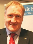 Lächelnder Mann mit rotblondem Haar in Anzug und roter Krawatte, mit politischem Banner im Hintergrund.