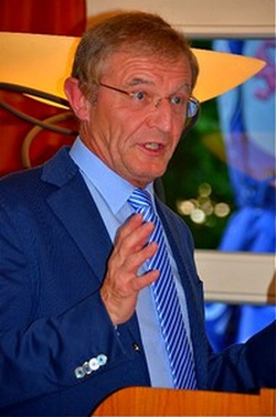 Ein Mann im blauen Anzug mit gestreifter Krawatte spricht hinter einem Holzpult und gestikuliert mit der Hand.
